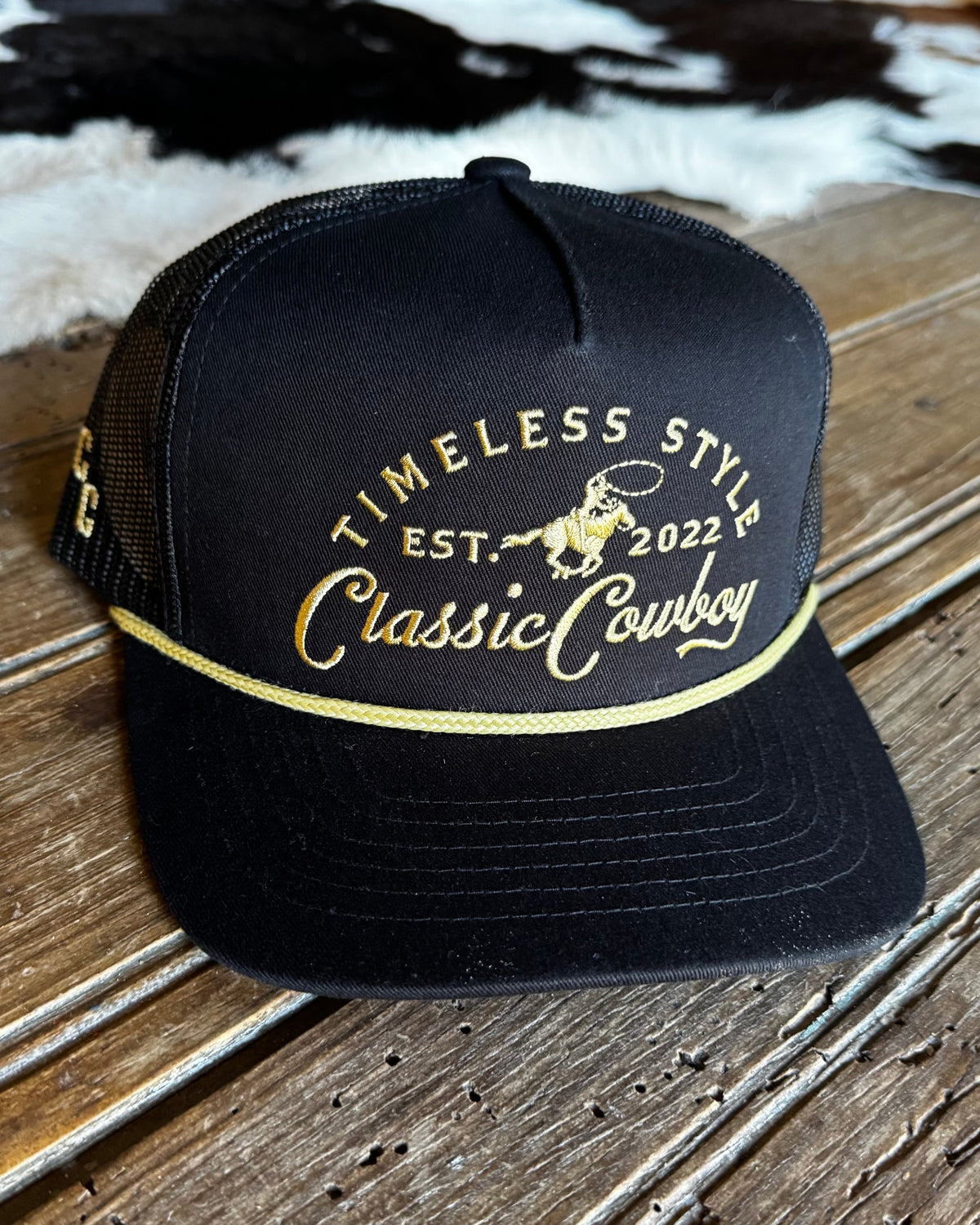 A black trucker hat with gold embroidered text reading 'TIMELESS STYLE EST. 2022 Classic Cowboy' and a cowboy graphic on the front panel, resting on a rustic wooden surface.