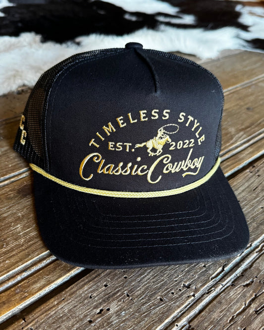 A black trucker hat with gold embroidered text reading 'TIMELESS STYLE EST. 2022 Classic Cowboy' and a cowboy graphic on the front panel, resting on a rustic wooden surface.