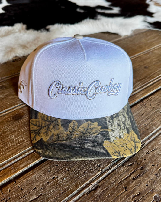 A structured baseball cap with a white crown and a camouflage-patterned visor, featuring the words 'Classic Cowboy' embroidered in white script on the front. The hat is displayed on a rustic wooden surface with a cowhide rug in the background.