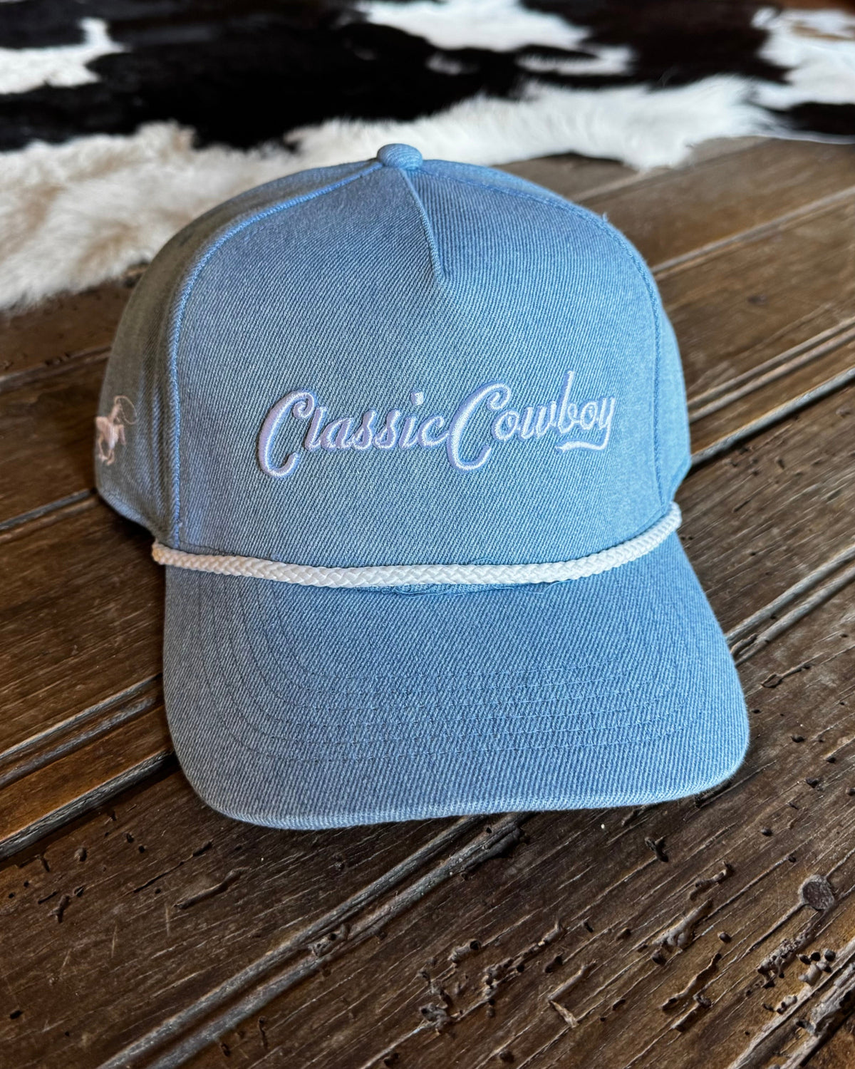 A light blue denim-style baseball cap with 'Classic Cowboy' embroidered in white script on the front, resting on a rustic wooden surface with a cowhide rug in the background.