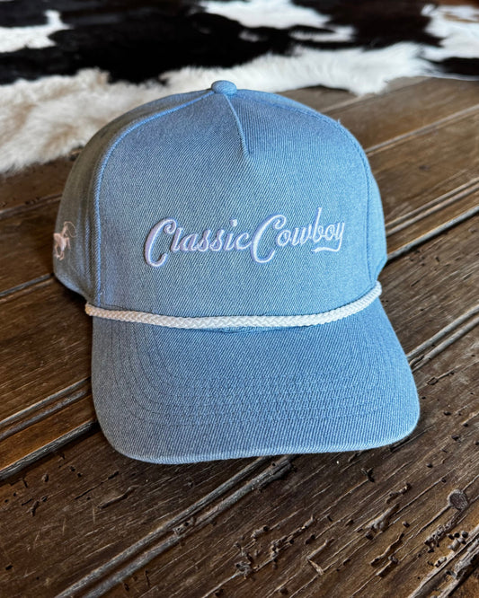 A light blue denim-style baseball cap with 'Classic Cowboy' embroidered in white script on the front, resting on a rustic wooden surface with a cowhide rug in the background.