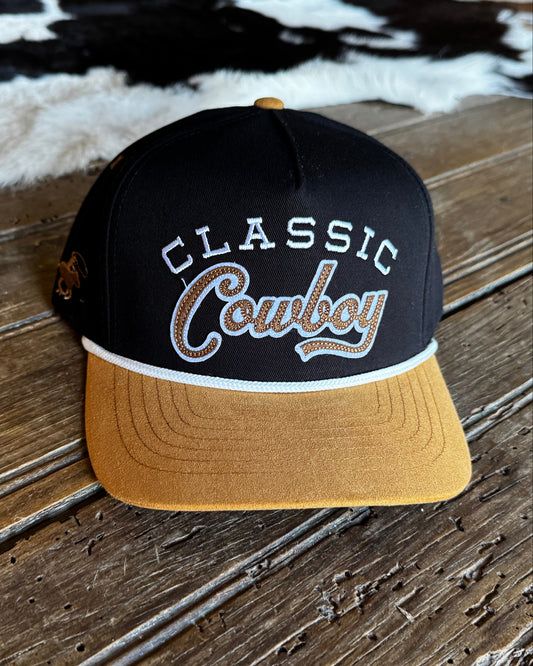 A black and gold snapback cap with 'CLASSIC Cowboy' embroidered on the front, resting on a rustic wooden table with a cowhide rug in the background.