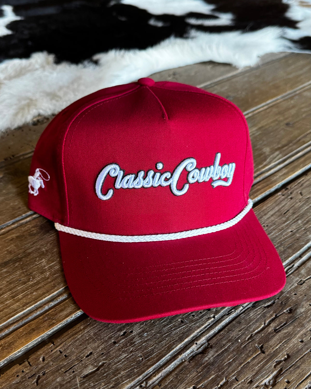 A red baseball cap with 'Classic Cowboy' embroidered in white script on the front, resting on a rustic wooden surface with a cowhide rug in the background.