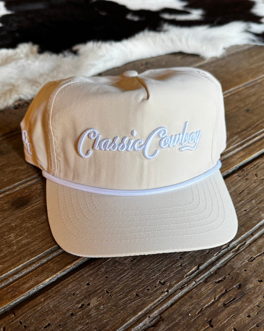 A beige baseball cap with 'Classic Cowboy' embroidered in white script on the front, resting on a rustic wooden surface with a cowhide rug in the background.