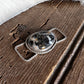 A close-up of a silver-toned metal accessory with an oval black-and-white marbled stone centerpiece, shown against a rustic brown wood background with white fur on the right.