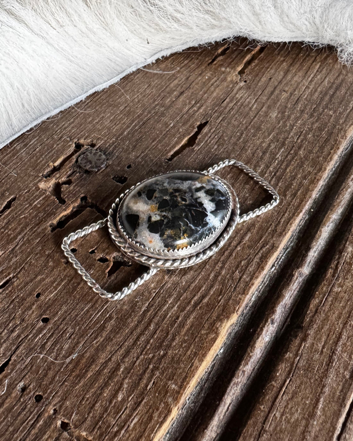 A close-up of a silver-toned metal accessory with an oval black-and-white marbled stone centerpiece, shown against a rustic brown wood background with white fur on the right.