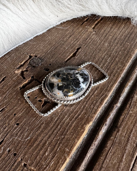 A close-up of a silver-toned metal accessory with an oval black-and-white marbled stone centerpiece, shown against a rustic brown wood background with white fur on the right.