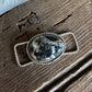 A silver-toned braided belt buckle with an oval cabochon stone featuring black, white, and gray marbled patterns, resting on a rustic dark wood surface.