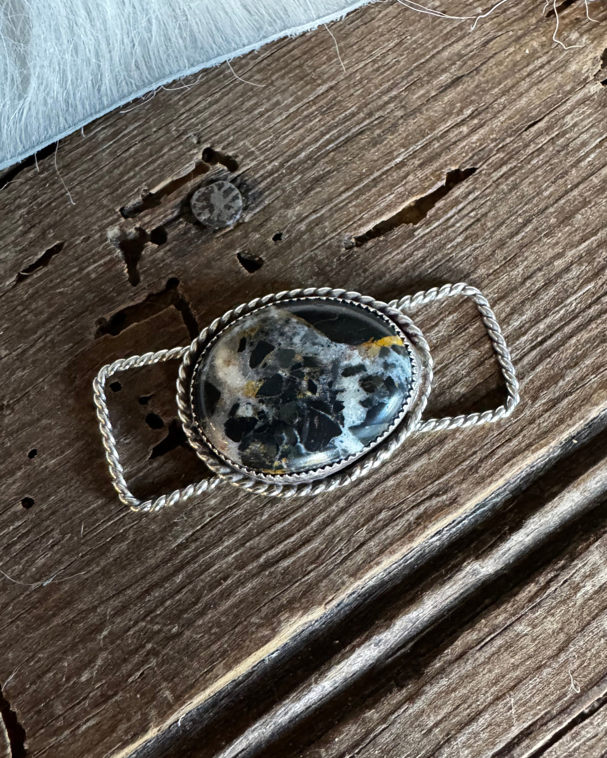 A silver-toned braided belt buckle with an oval cabochon stone featuring black, white, and gray marbled patterns, resting on a rustic dark wood surface.