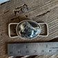 A silver-toned oval pendant with a black, white, and gray marbled stone, displayed next to a stainless steel ruler on a rustic wooden surface.