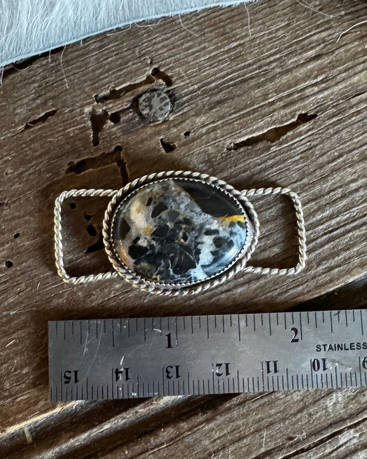 A silver-toned oval pendant with a black, white, and gray marbled stone, displayed next to a stainless steel ruler on a rustic wooden surface.