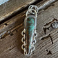 A handmade sterling silver pendant featuring a central turquoise stone, displayed on a rustic dark wood surface.