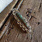 A handmade sterling silver pendant with a green turquoise stone, resting on a rustic wooden surface next to white fur.