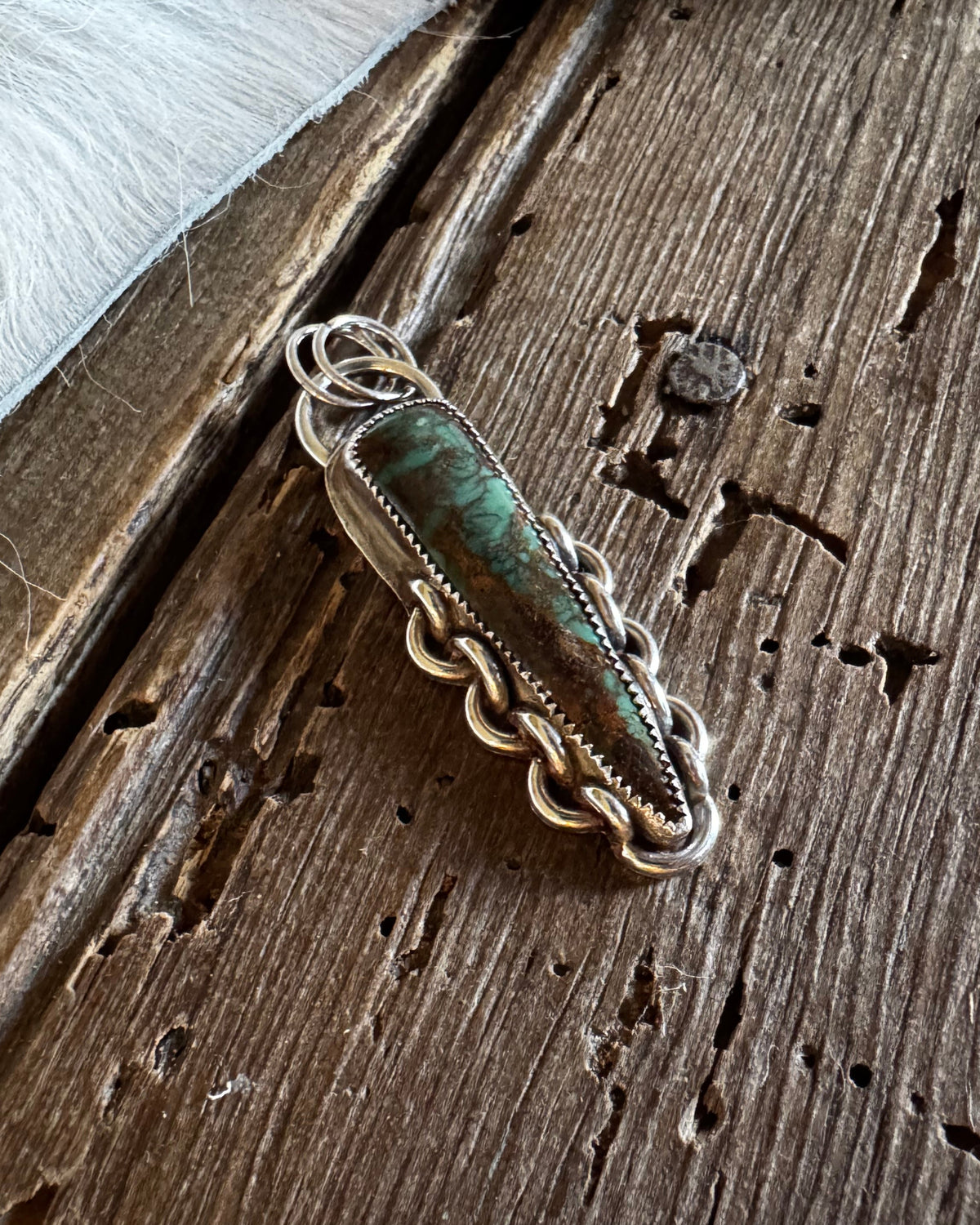 A handmade sterling silver pendant with a green turquoise stone, resting on a rustic wooden surface next to white fur.