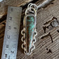 A handmade sterling silver pendant with a green turquoise stone, shown next to a stainless steel ruler on a rustic wooden surface.