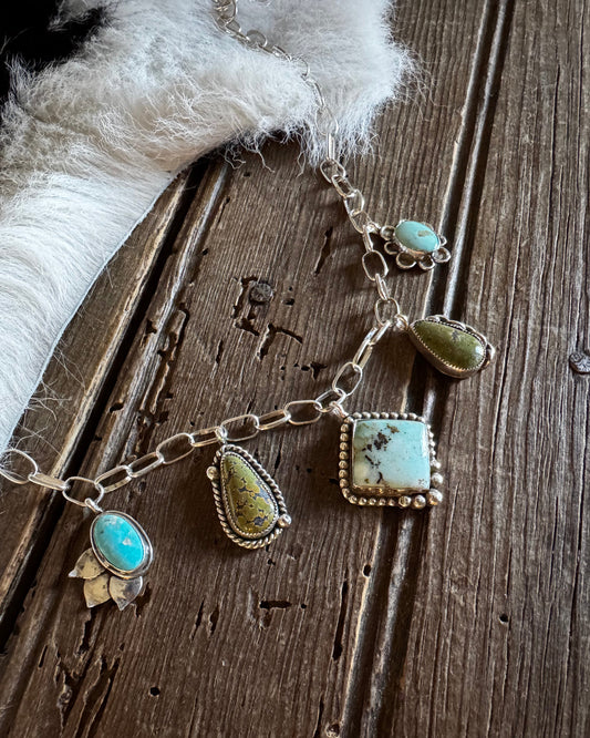 A sterling silver chain bracelet featuring multiple turquoise and green stone charms, displayed on a rustic dark wood surface next to a white faux fur accent.