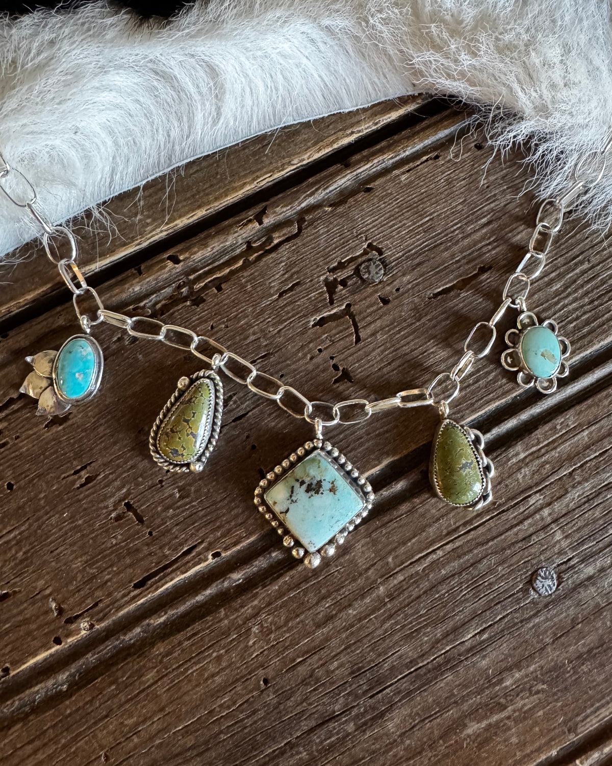 A silver chain bracelet with multiple turquoise and green stone charms, styled on a rustic dark wood background next to a white faux fur trim.