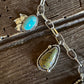 A close-up of a silver-toned jewelry necklace laid on a rustic dark wood surface, featuring multiple stone pendants including turquoise, greenish-yellow marbled stone, and a pale blue square stone with dark matrix.