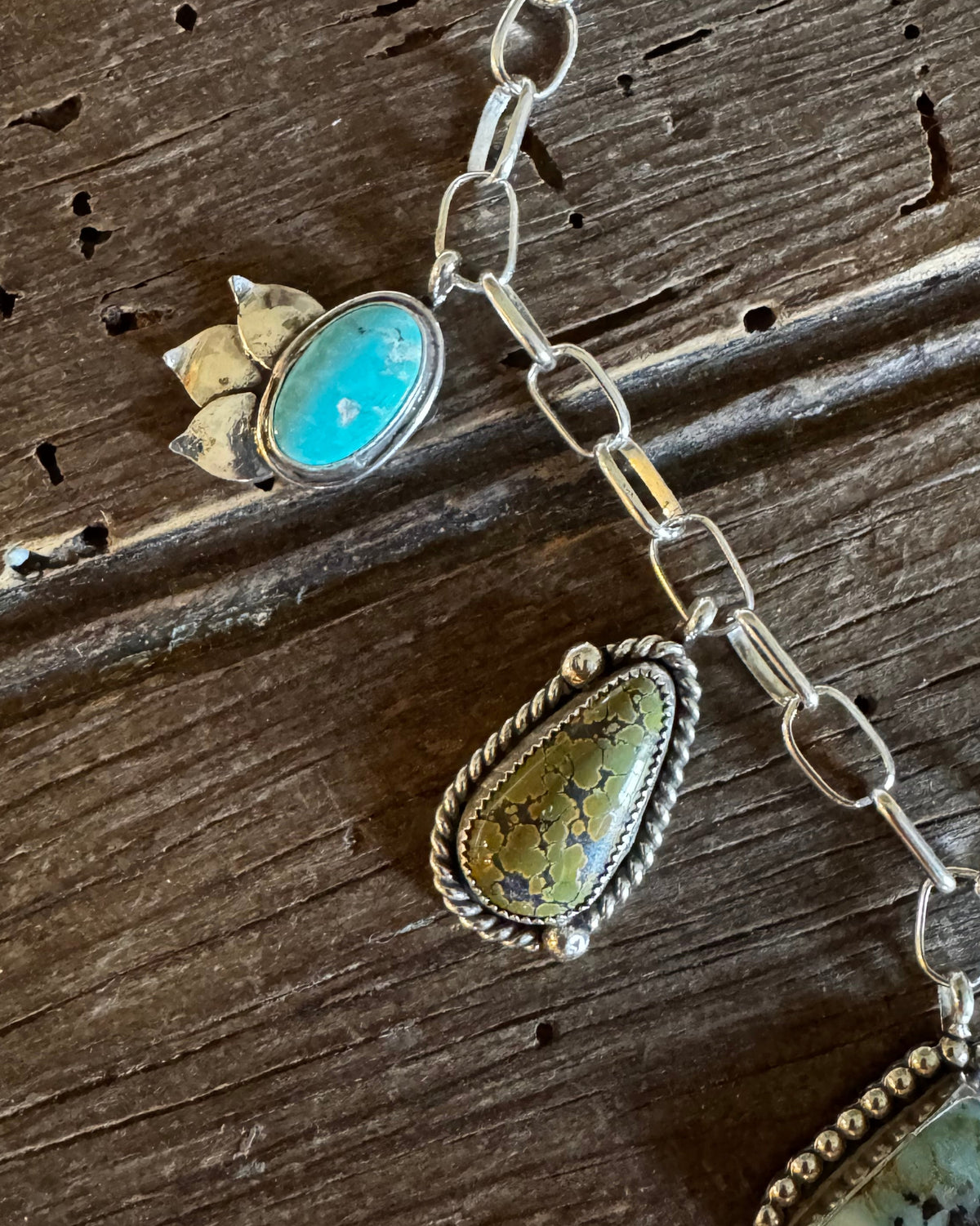 A close-up of a silver-toned jewelry necklace laid on a rustic dark wood surface, featuring multiple stone pendants including turquoise, greenish-yellow marbled stone, and a pale blue square stone with dark matrix.