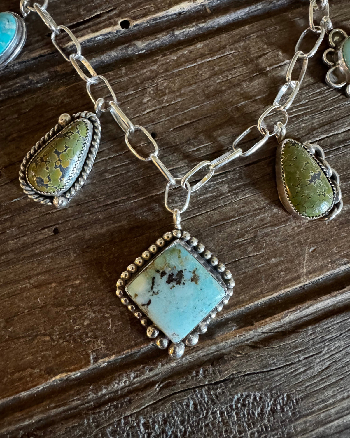 A silver-toned chain necklace with multiple turquoise stone pendants, including a large diamond-shaped pendant in the center and two smaller teardrop pendants on either side, displayed on a rustic dark wood surface.