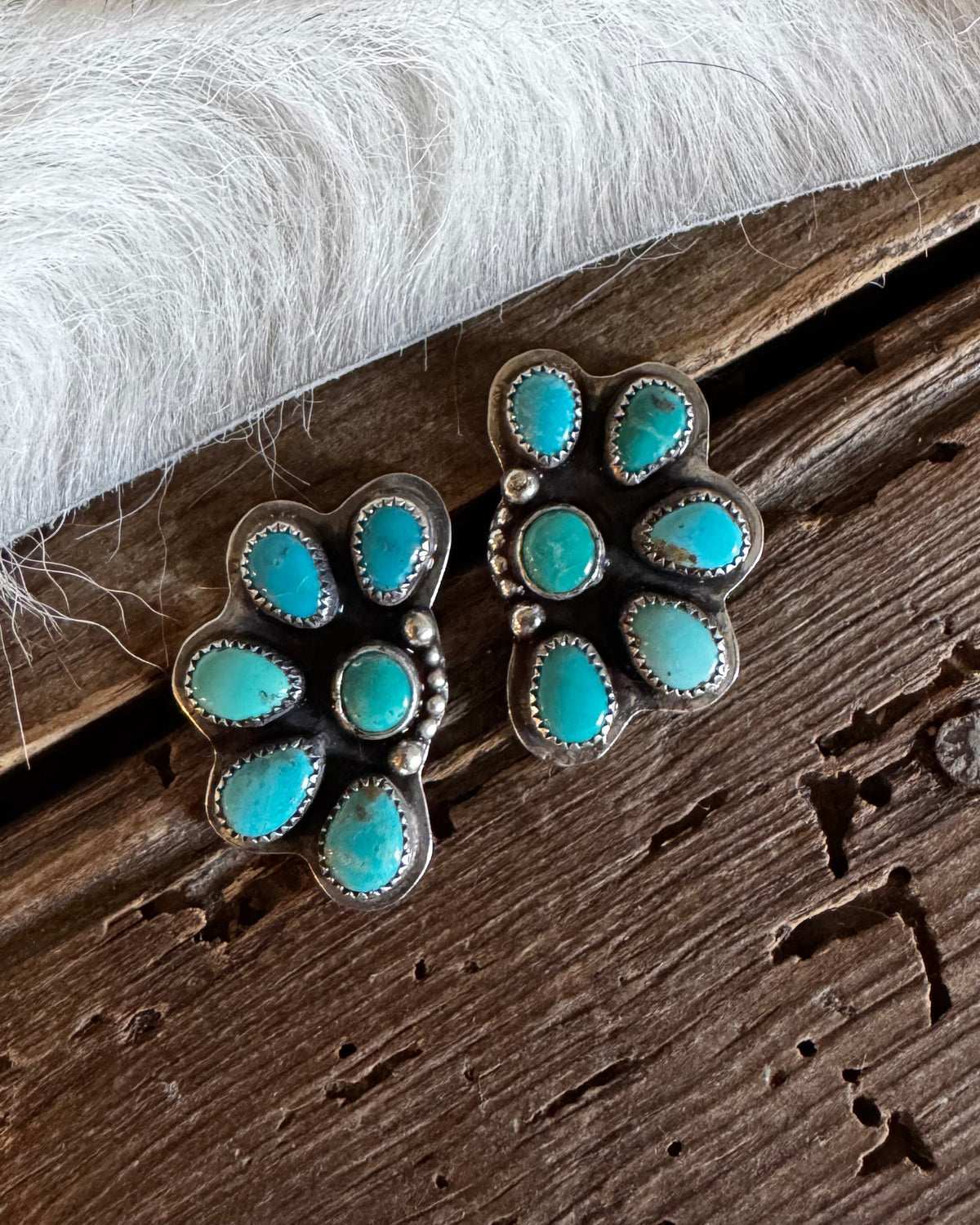 A pair of silver-toned floral stud earrings with turquoise stones, displayed on a rustic dark wood surface next to white faux fur.