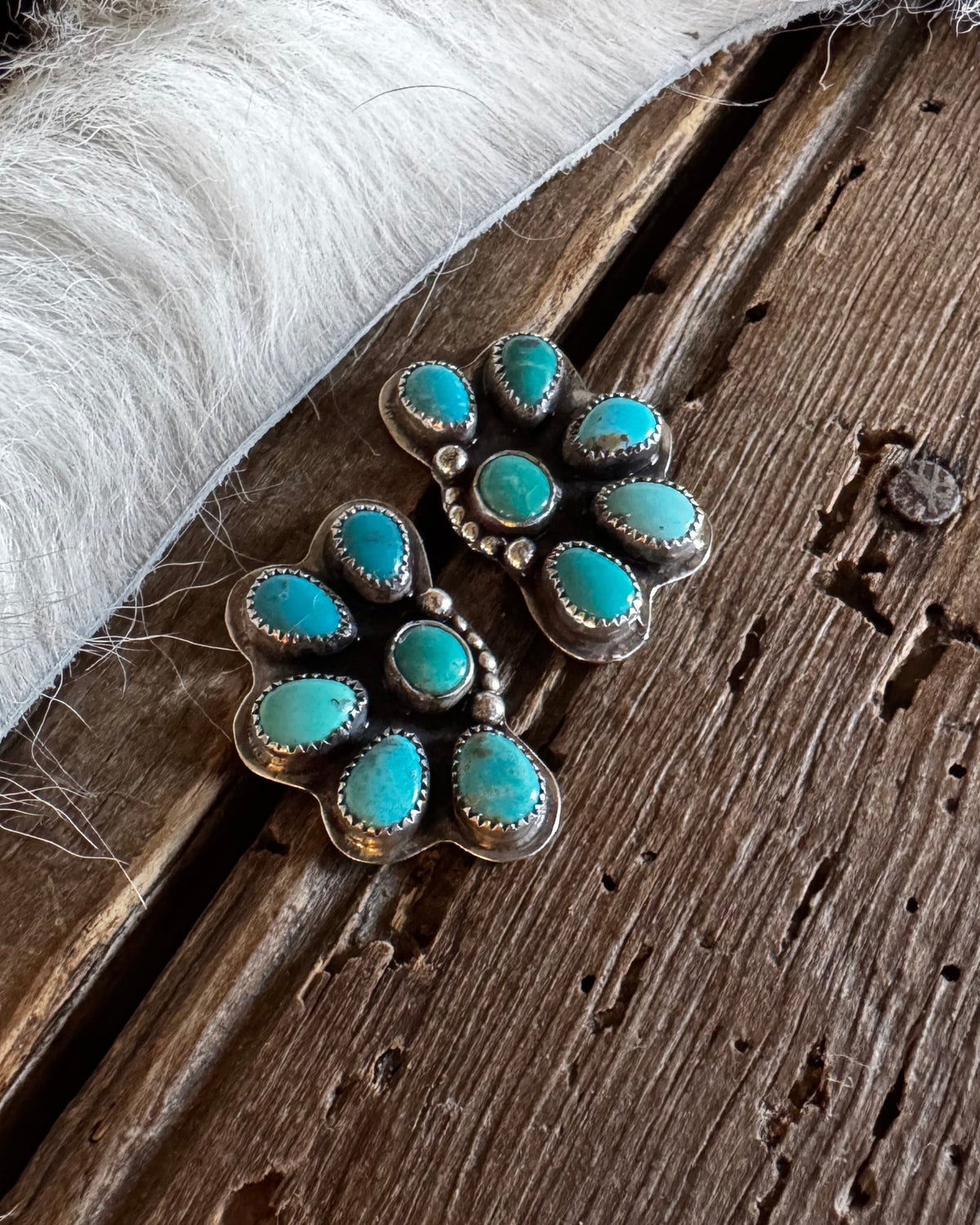 A pair of silver-toned floral stud earrings with turquoise stones, resting on a rustic dark wood surface next to a white faux fur trim.