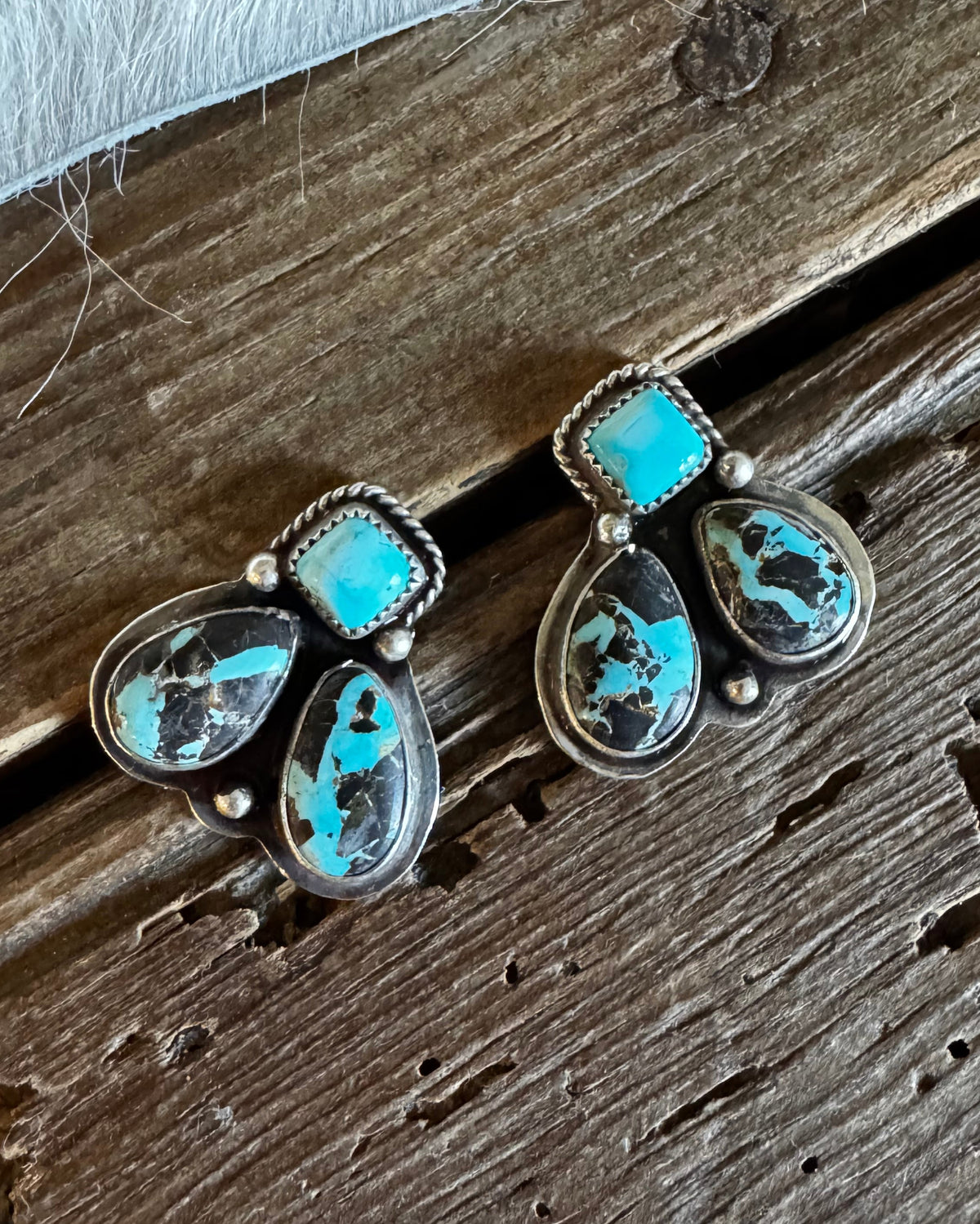 A pair of silver-toned drop earrings with rectangular turquoise stones and teardrop-shaped turquoise and black marbled stones, displayed on a rustic wooden surface.