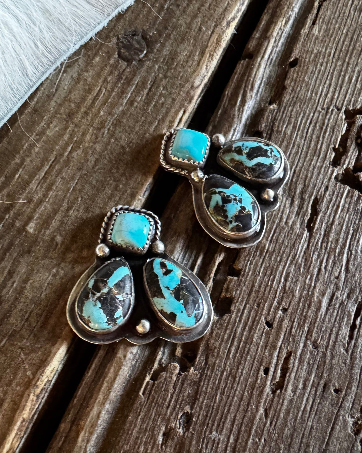 A pair of silver dangle earrings with turquoise and black marbled stones, displayed on a rustic wooden surface next to a white faux fur trim.