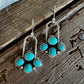 A pair of silver dangle earrings with turquoise stone clusters, displayed on a rustic dark wood surface.