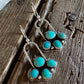 A pair of silver-toned dangle earrings with five turquoise stones each, displayed on a rustic dark wood surface next to a white faux fur trim.