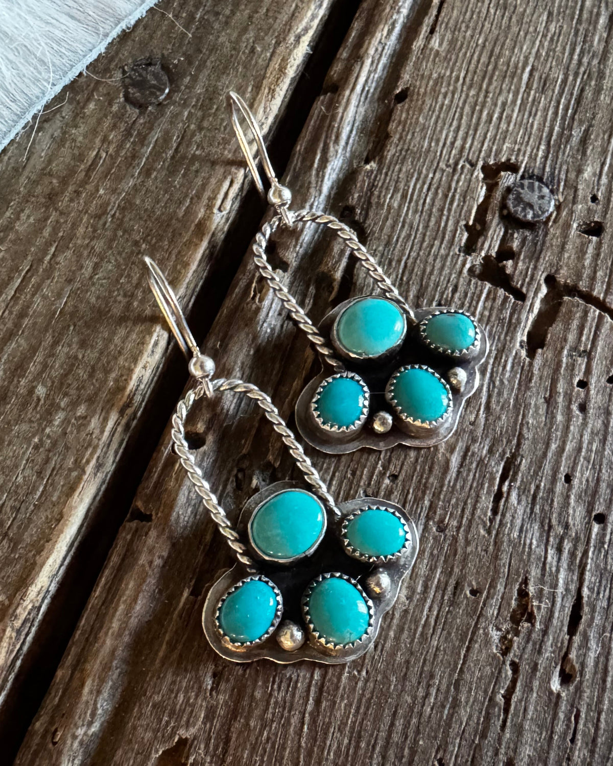 A pair of silver-toned dangle earrings with five turquoise stones each, displayed on a rustic dark wood surface next to a white faux fur trim.