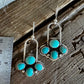 A pair of silver dangle earrings with turquoise stones, displayed on a rustic wooden surface next to a metal ruler for scale.