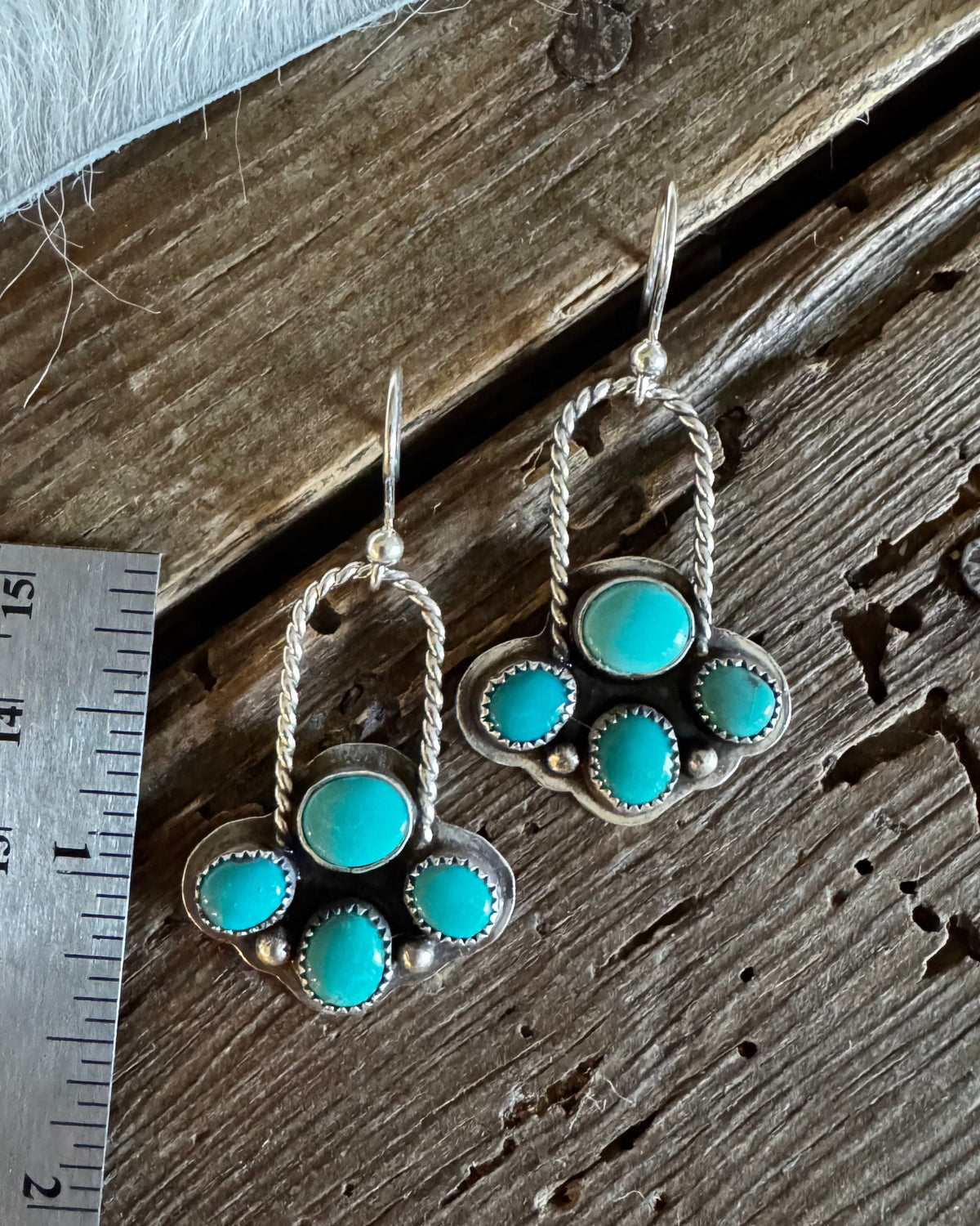 A pair of silver dangle earrings with turquoise stones, displayed on a rustic wooden surface next to a metal ruler for scale.