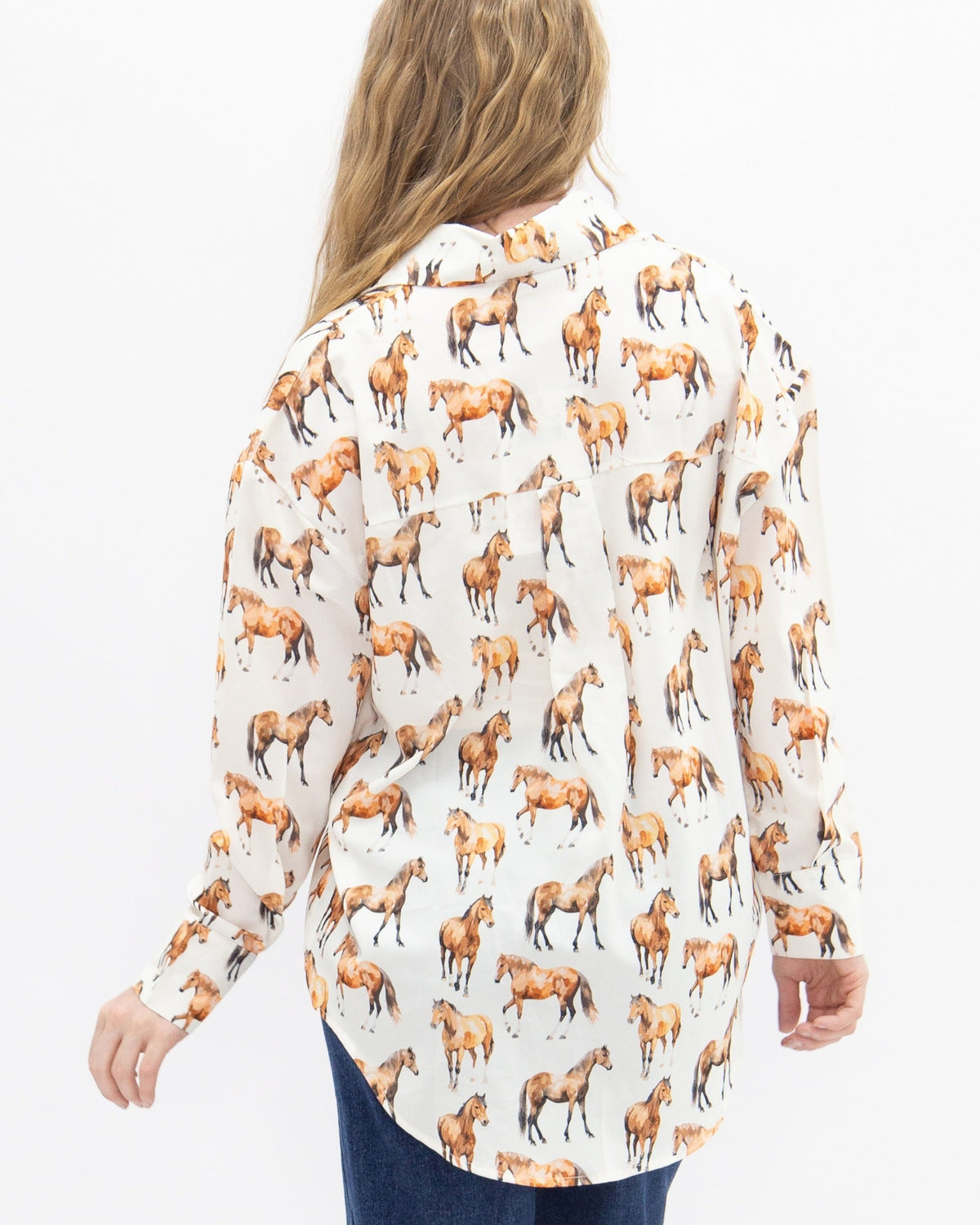 A woman viewed from behind wearing a long-sleeve button-up shirt with an all-over print of brown and black horses on a white background, paired with dark blue jeans against a plain white studio background.