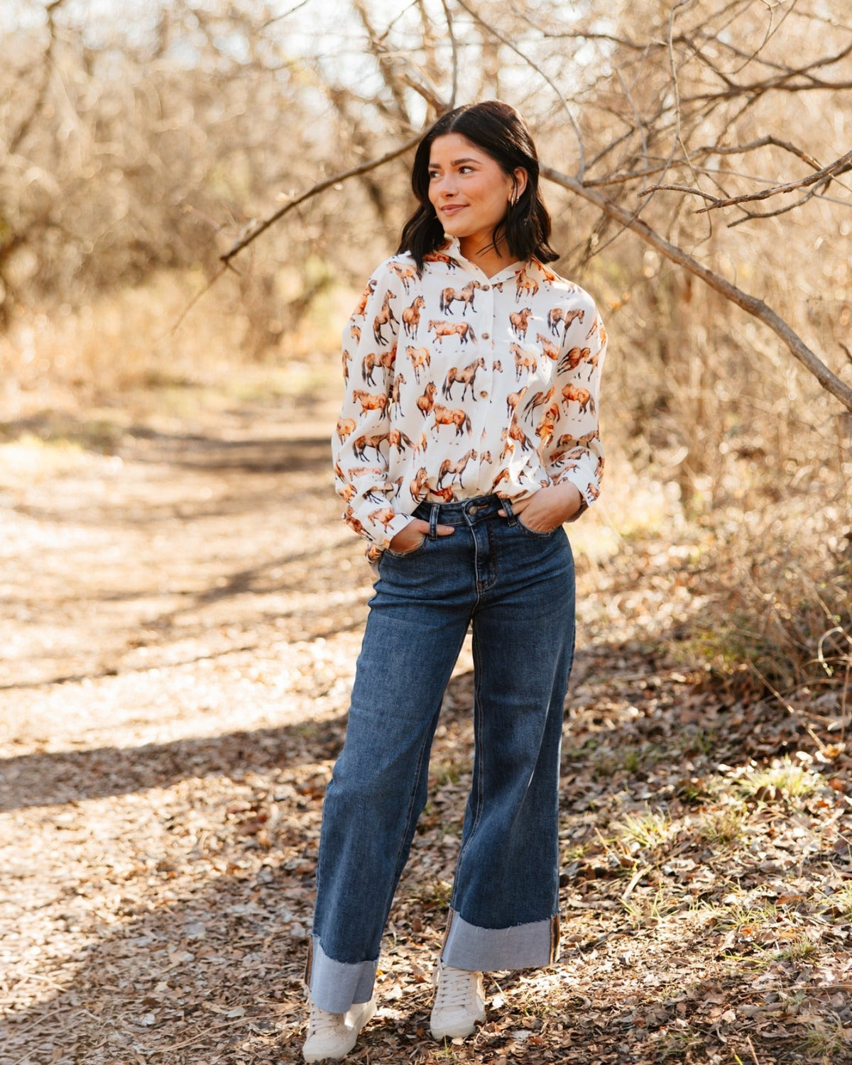 Woman standing in a forest wearing a patterned blouse and jeans.