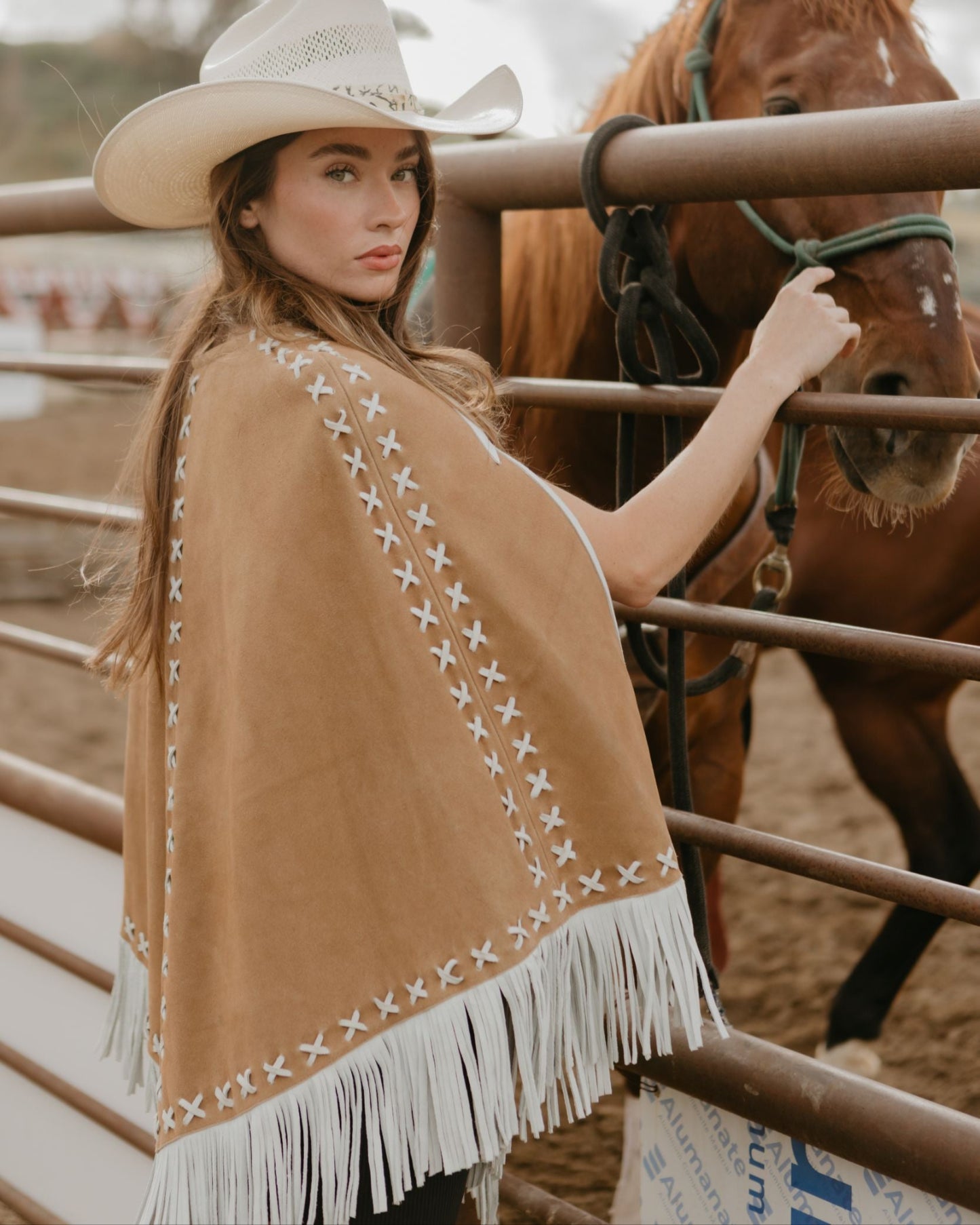 Woman wearing a brown suede leather cape standing by a fence with a horse 