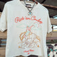An off-white short-sleeve graphic t-shirt hanging on a wooden hanger. The shirt features a western-themed graphic with the text 'Ride 'em Cowboy' in red, an illustration of a cowboy on a bucking horse, and 'American Ranch 1865' printed below.