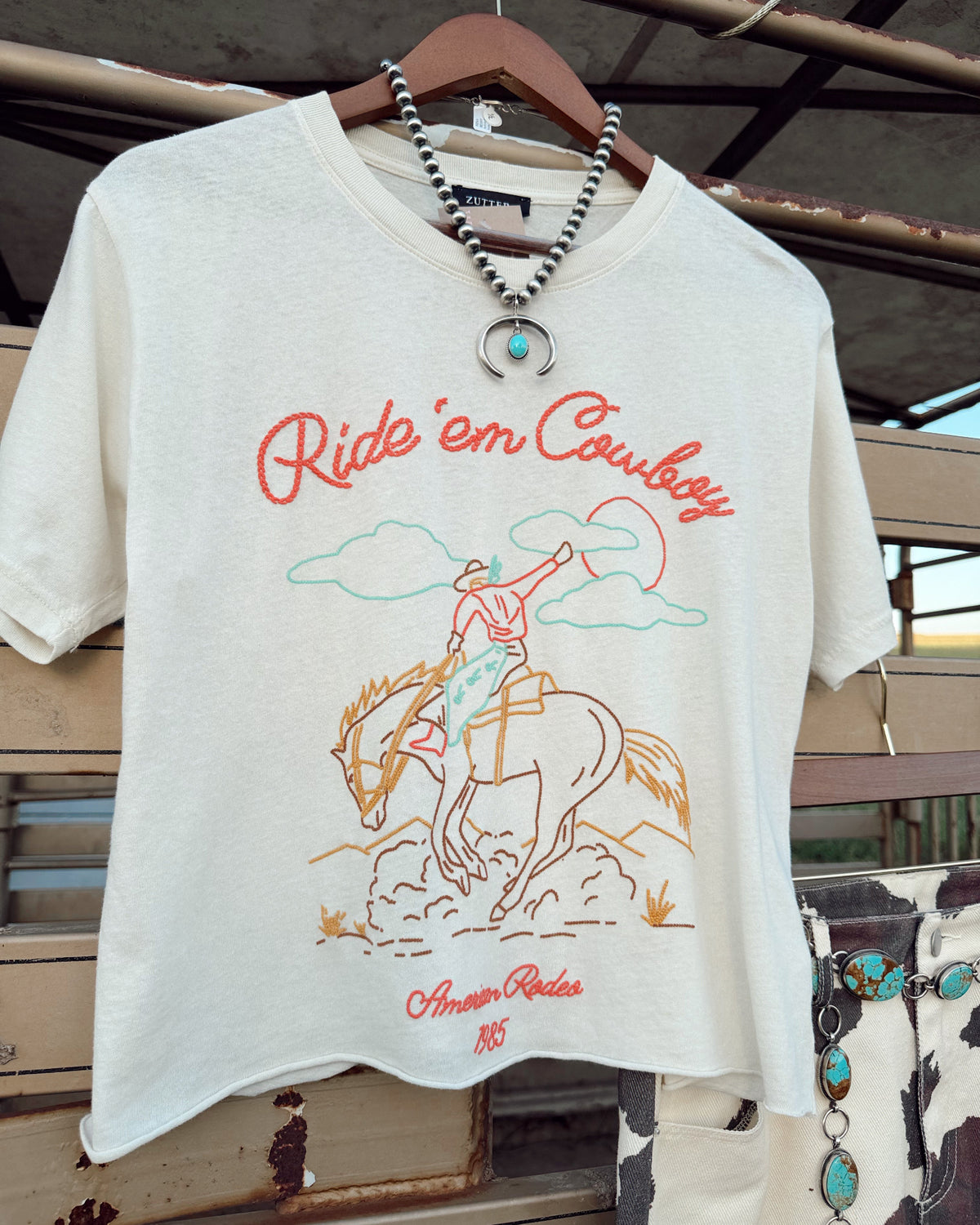 An off-white short-sleeve graphic t-shirt hanging on a wooden hanger. The shirt features a western-themed graphic with the text 'Ride 'em Cowboy' in red, an illustration of a cowboy on a bucking horse, and 'American Ranch 1865' printed below.