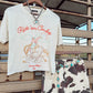A western-themed outfit set hanging on a wooden stand, featuring an off-white graphic t-shirt with 'Ride 'em Cowboy' text and an illustration, paired with matching cow-print shorts and turquoise bead accessories.