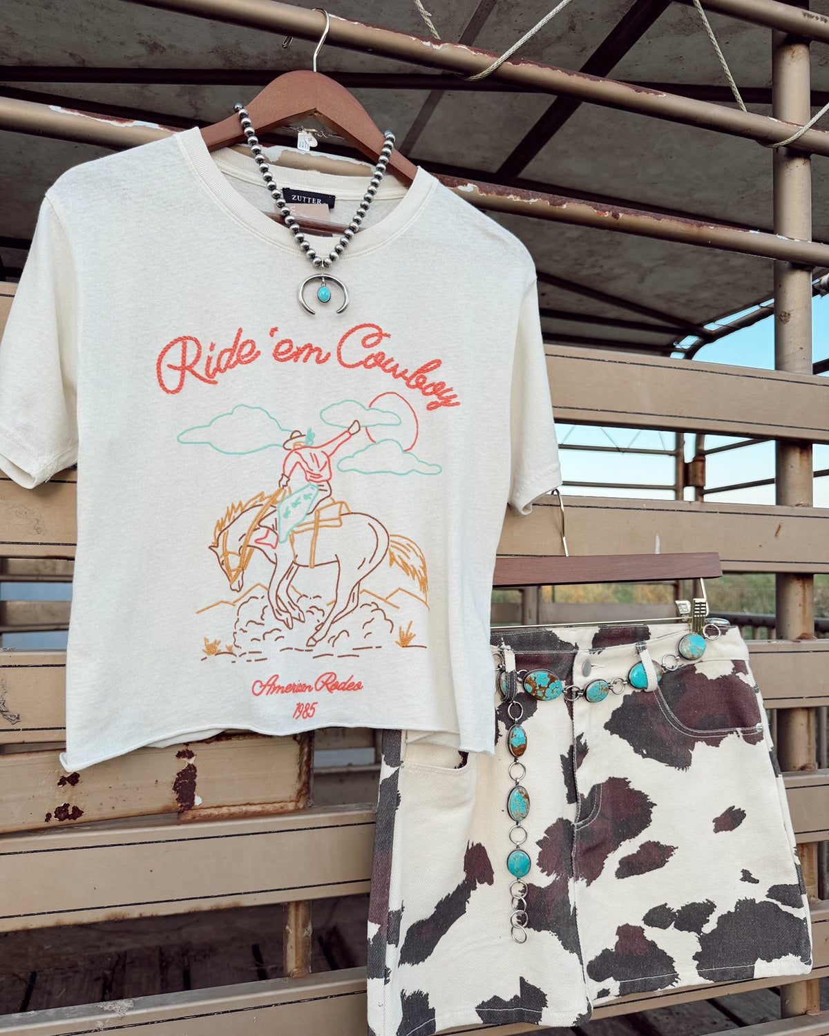 A western-themed outfit set hanging on a wooden stand, featuring an off-white graphic t-shirt with 'Ride 'em Cowboy' text and an illustration, paired with matching cow-print shorts and turquoise bead accessories.