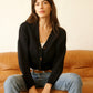 A woman sits on a tan leather sofa wearing a black knit cardigan with brown buttons over a white top and light blue jeans, against a textured off-white wall.