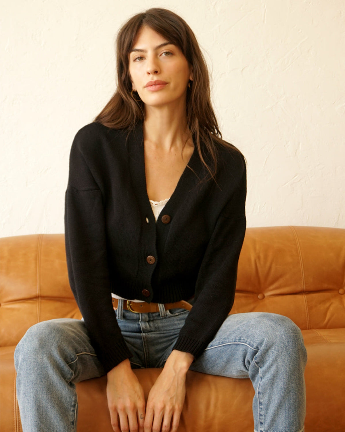 A woman sits on a tan leather sofa wearing a black knit cardigan with brown buttons over a white top and light blue jeans, against a textured off-white wall.