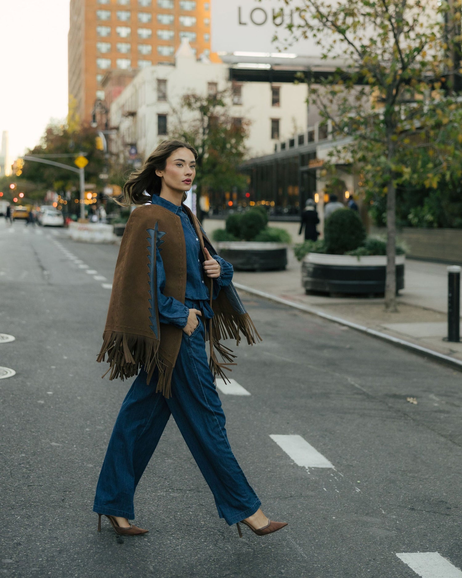 Woman walking on a city street with a brown fringed jacket and blue pants.