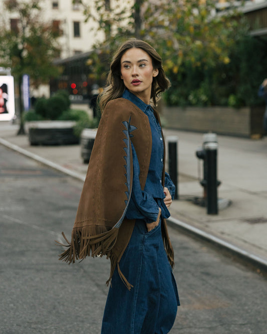 Woman walking on a city street wearing a brown fringed jacket and blue outfit.