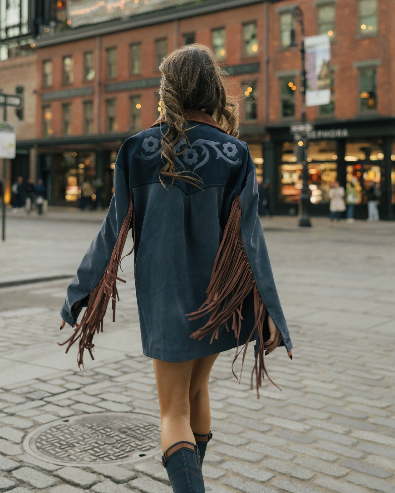 Person wearing a blue leather jacket with fringe walking on a city street.